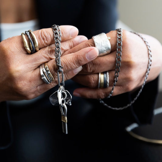 Close-up of hands wearing multiple silver and bronze rings and a talisman chain with various talisman pendants.