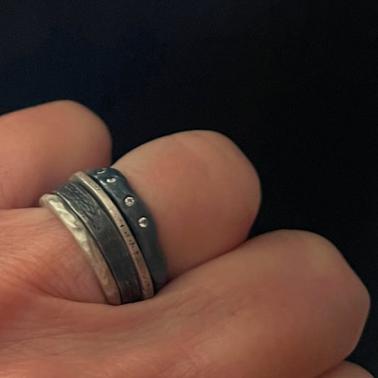 Close-up of a hand wearing a stack of silver and oxidized silver rings by Chikahisa Studio on a dark background
